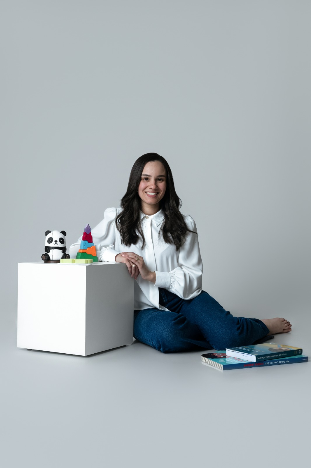 Yasmim Tagliatella sentada em estúdio ao lado de brinquedos e livros infantis.